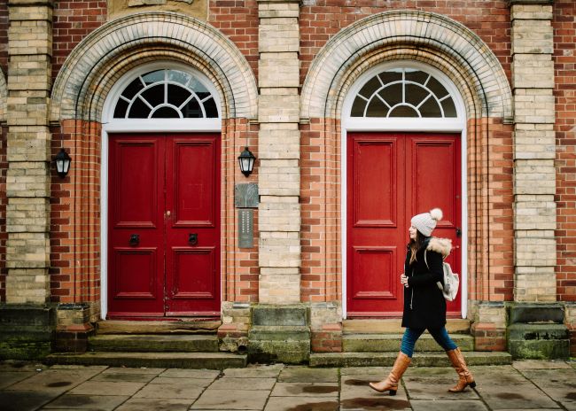 A woman wearing brown boots walks in front of two red doors